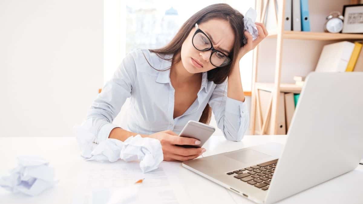woman wearing glasses at her computer looking frustrated at her phone.
