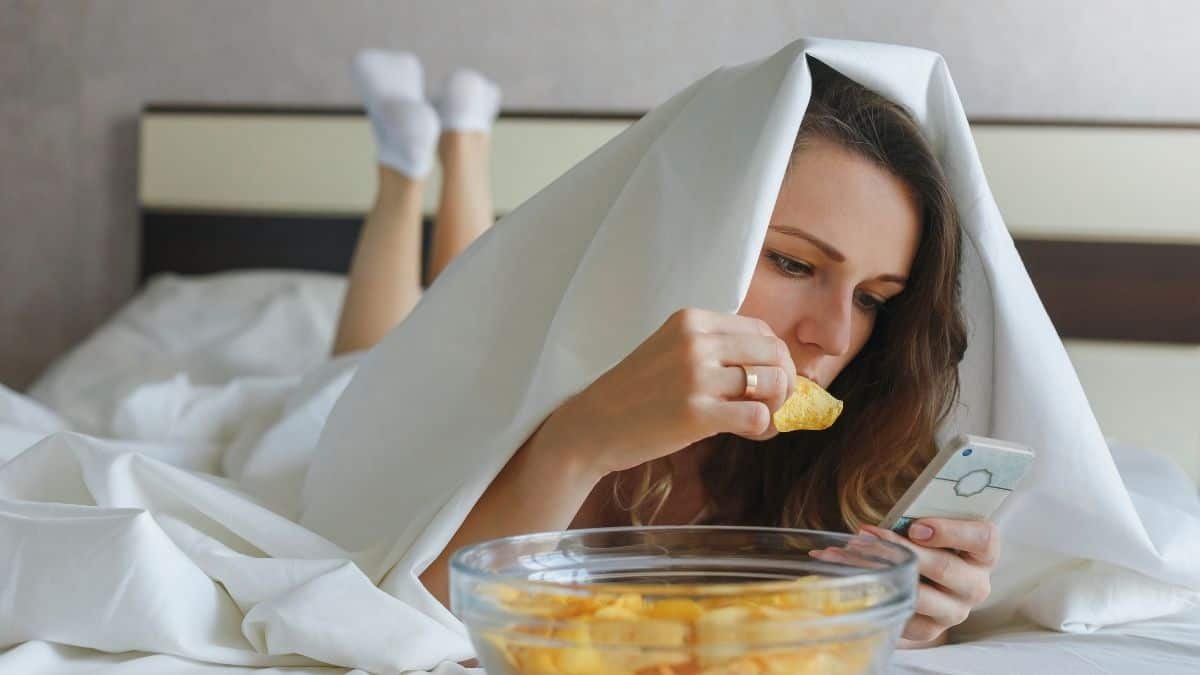 woman sitting in bed eating chips looking at her phone.