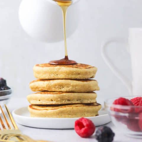 A stack of pancakes with maple syrup being poured on top.