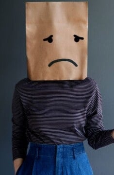 woman with paper bag on her head with a sad face drawn on.