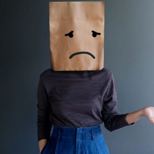 woman with paper bag on her head with a sad face drawn on.
