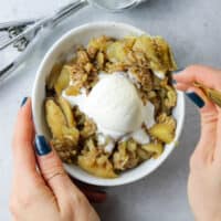 Two hands holding bowl of skillet apple crisp with ice cream and eating it.