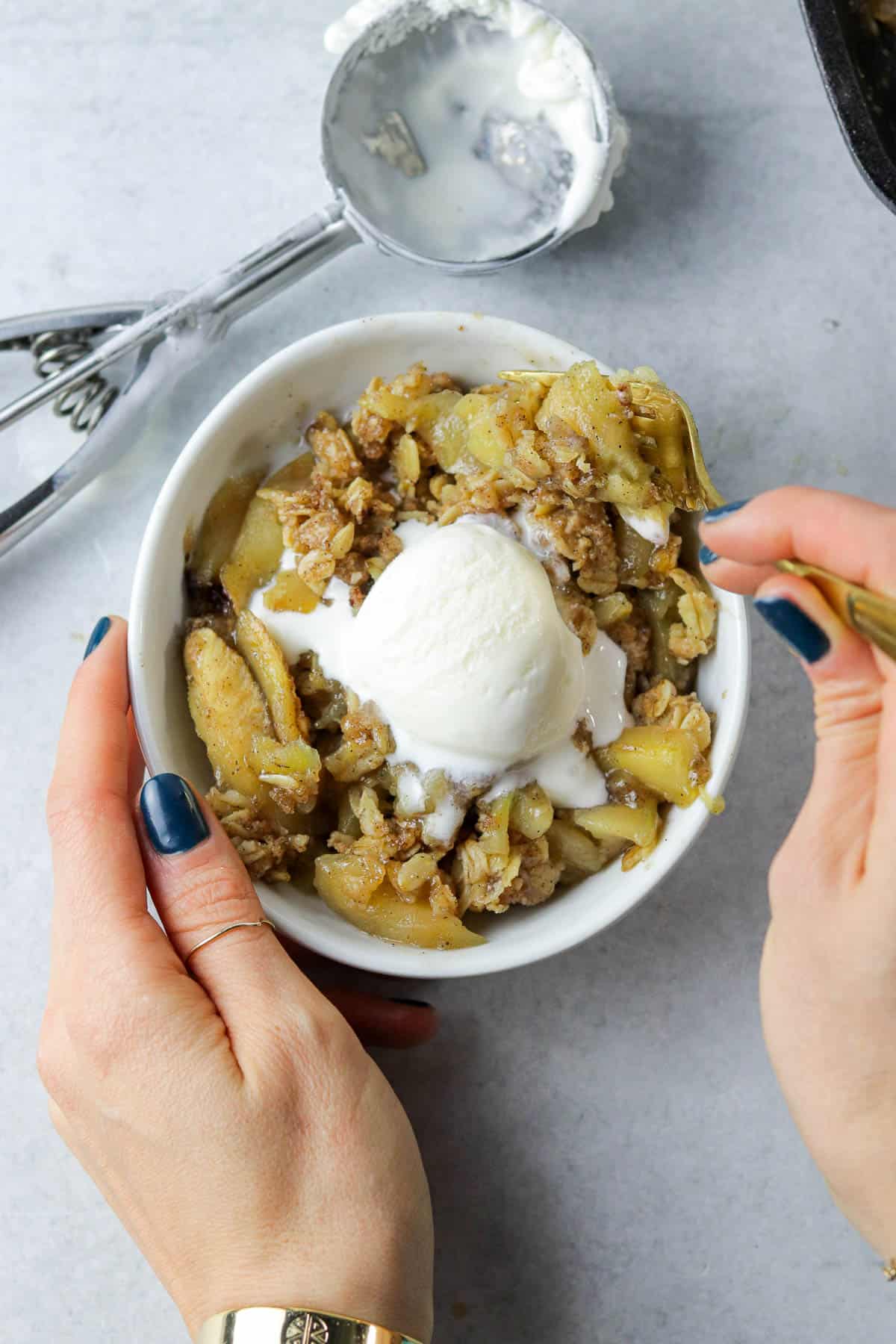 Two hands holding bowl of skillet apple crisp with ice cream and eating it.