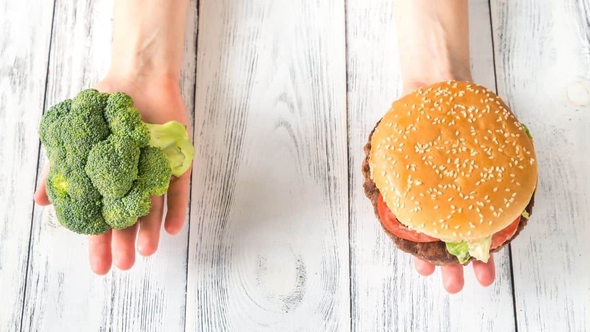 One hand holding a head of broccoli and one hand holding a burger over a wooden surface.