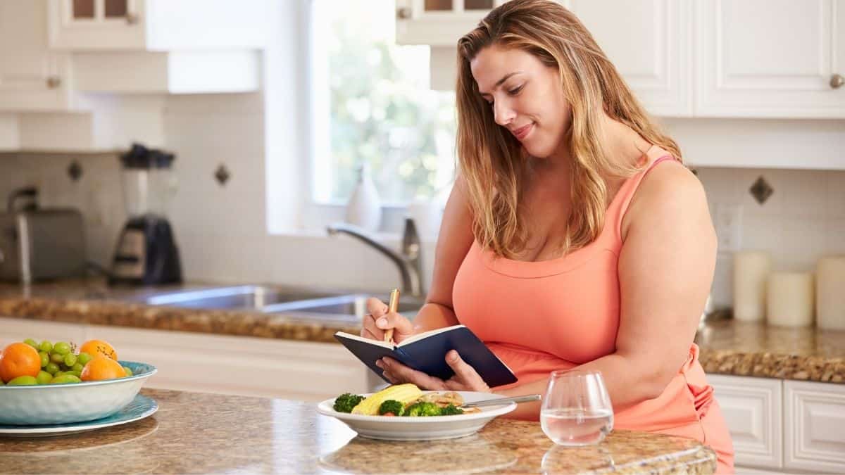 A woman preparing food in a kitchen, with notebook writing things down.