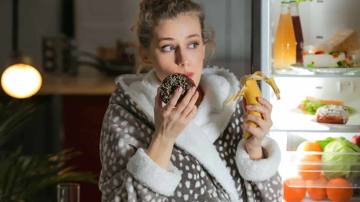 Woman in a bath robe eating a donut and a banana by the fridge.