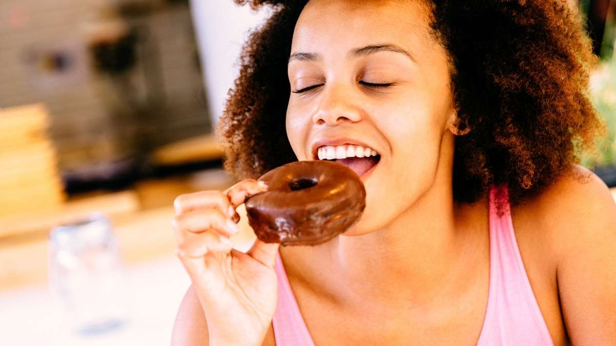 woman wearing a pink tank top eating a chocolate donut.