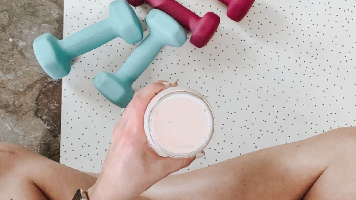 a woman holding a meal replacement drink while sitting on a yoga mat, next to some pink hand weights.