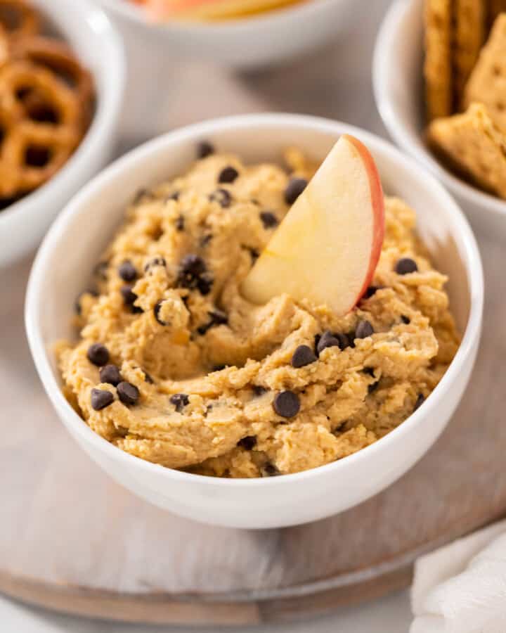 a bowl of Chocolate Chip Cookie Dough Hummus with a slice of apple dipped in and pretzels, sliced apples and graham crackers in the background.