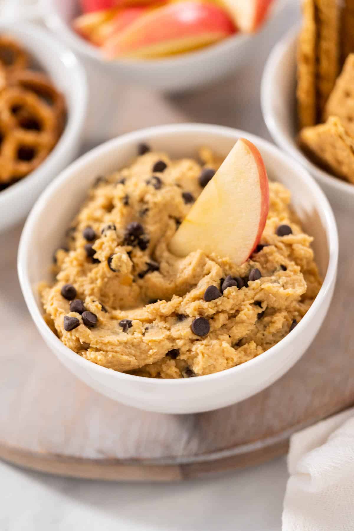 a bowl of Chocolate Chip Cookie Dough Hummus with a slice of apple dipped in and pretzels, sliced apples and graham crackers in the background.