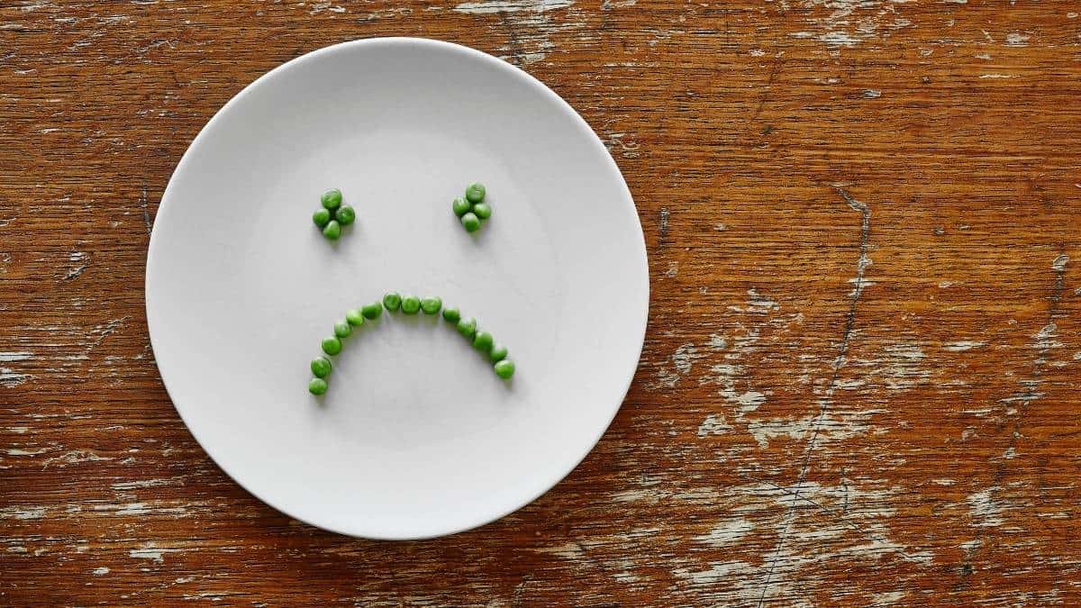 a white plate with a sad face spelled in peas on a wood table.