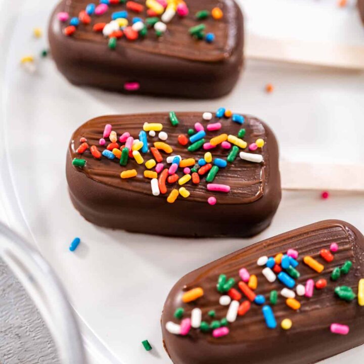 four chocolate covered cakesicles on a white plate with vibrant sprinkles on top.