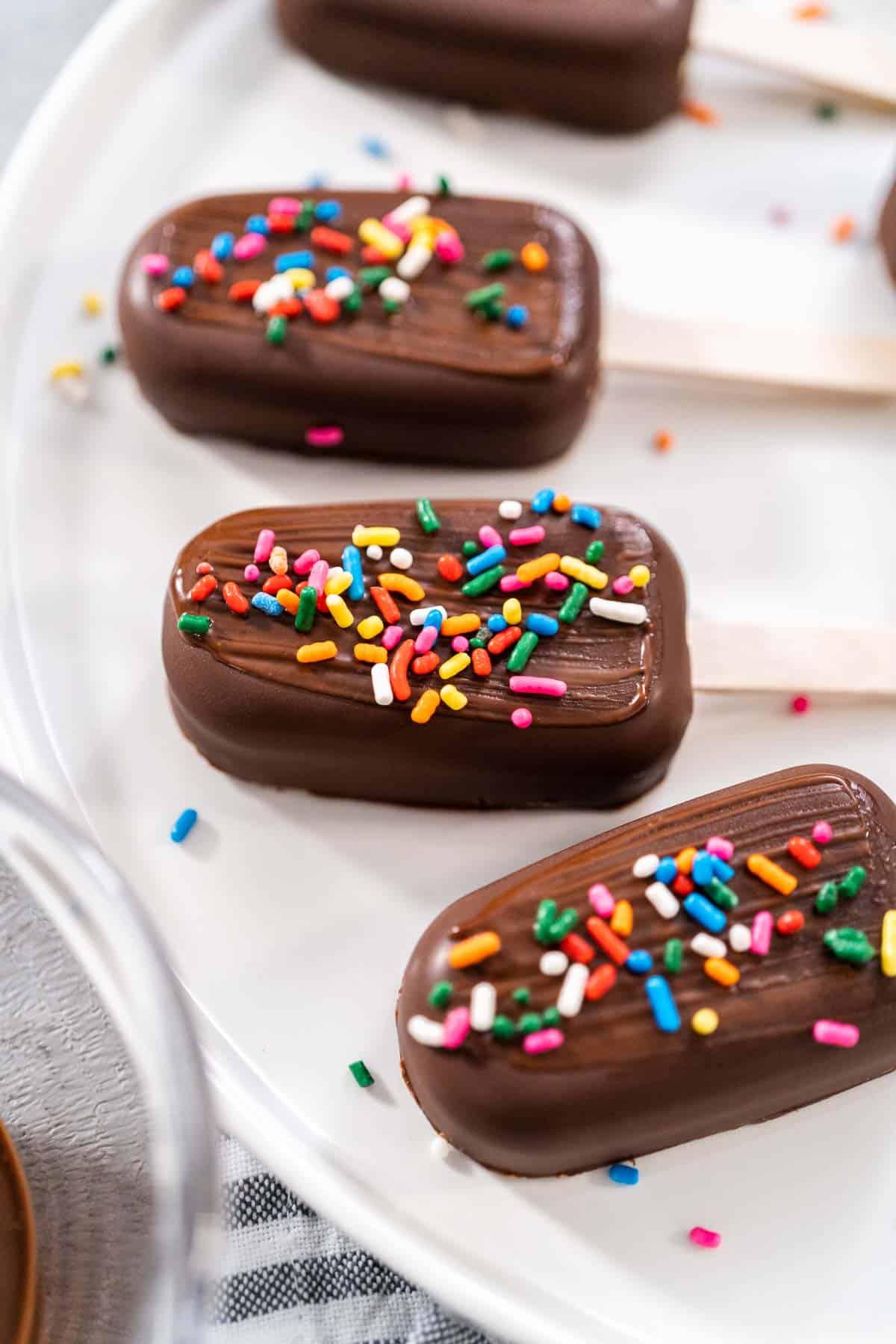 four chocolate covered cakesicles on a white plate with vibrant sprinkles on top.