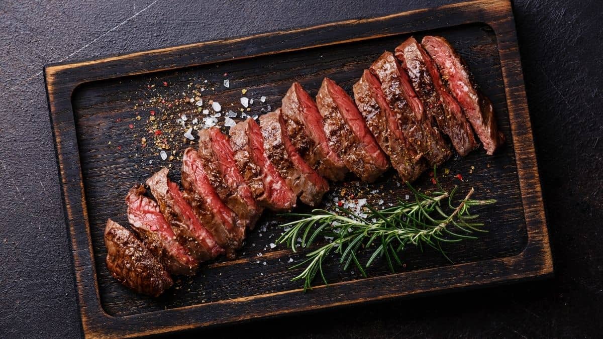 sliced steak with rosemary sprigs on the side on a wooden serving platter, an example of food you can eat on the slow carb diet.