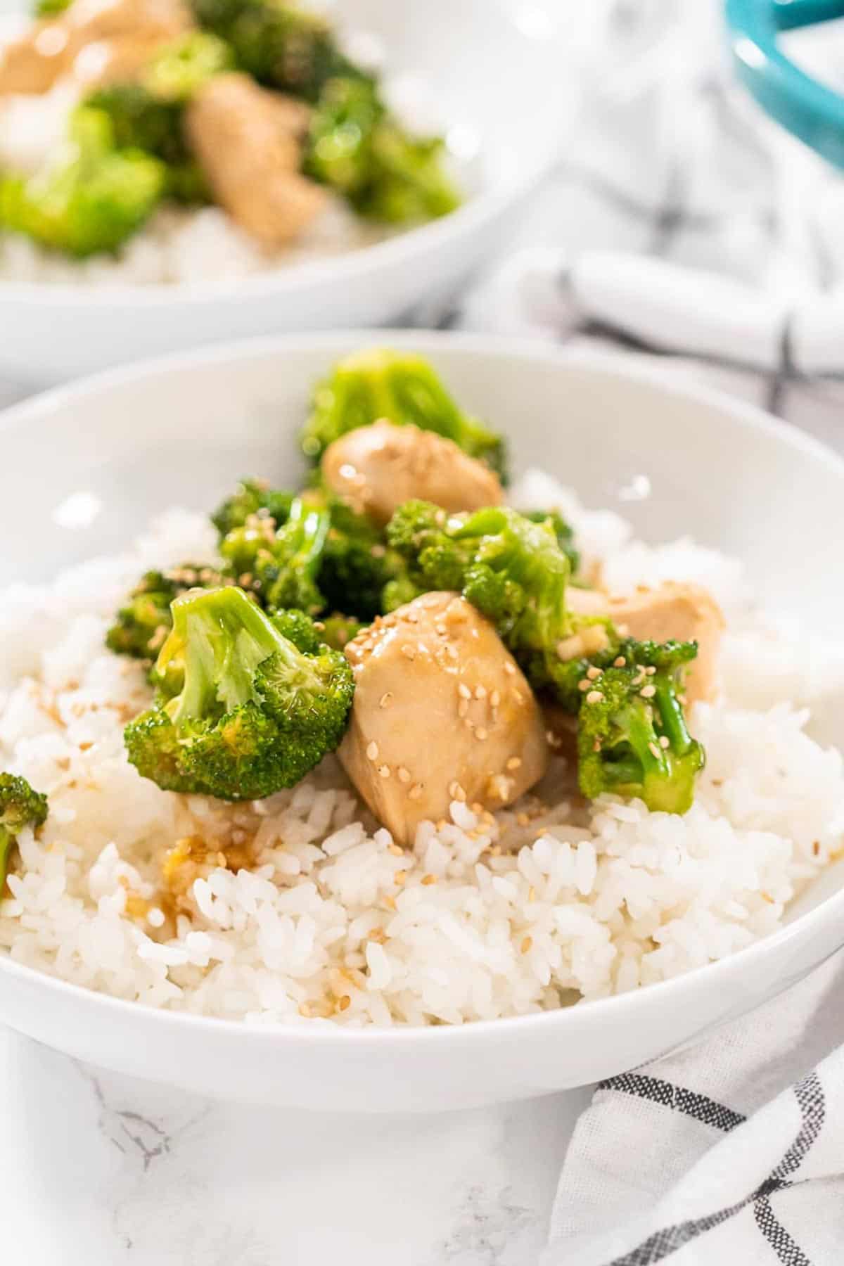A bowl of white rice and Easy Broccoli Chicken Stir Fry in a white bowl sitting on a white and grey kitchen towel.