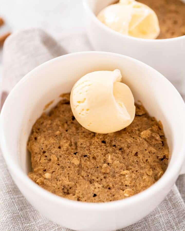 A close up of oatmeal apple mug cake in a white ceramic mug with a scoop of vanilla ice cream on top. A second mug sits in the background.