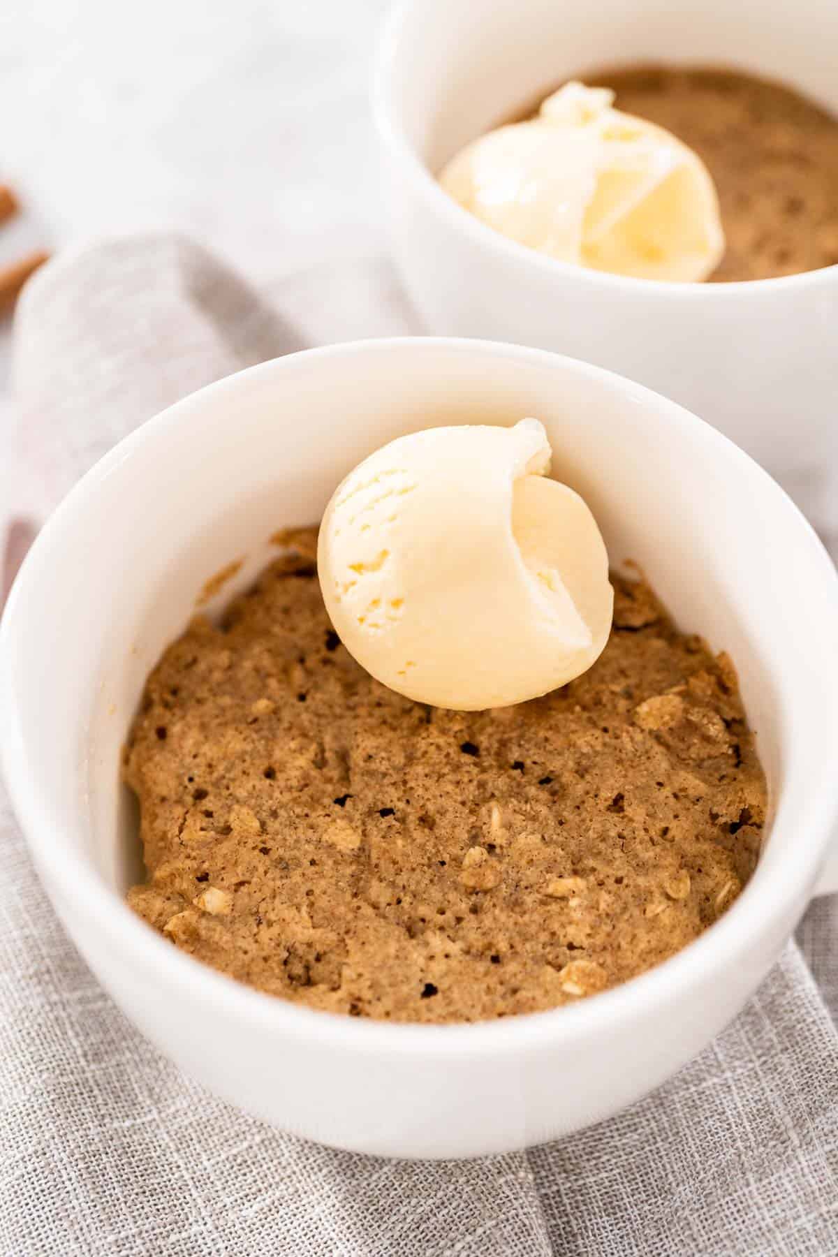A close up of oatmeal apple mug cake in a white ceramic mug with a scoop of vanilla ice cream on top. A second mug sits in the background.