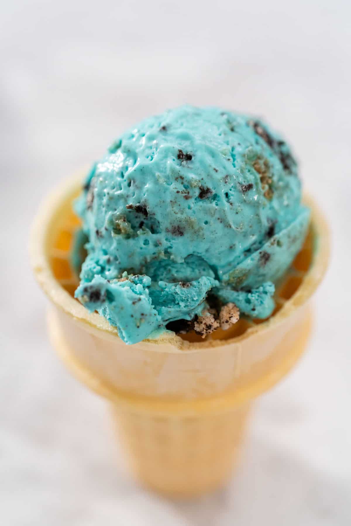 A scoop of Cookie Monster Ice Cream in a traditional ice cream cone on a grey and white countertop background.