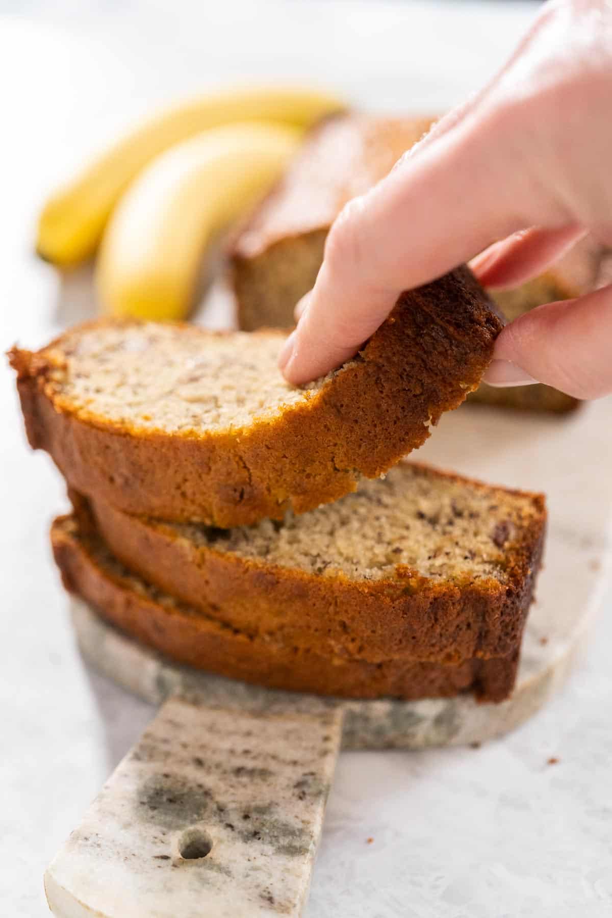 A stack of three pieces of one bowl banana bread with a woman's hand taking the top piece and two bananas and rest of loaf in the background.