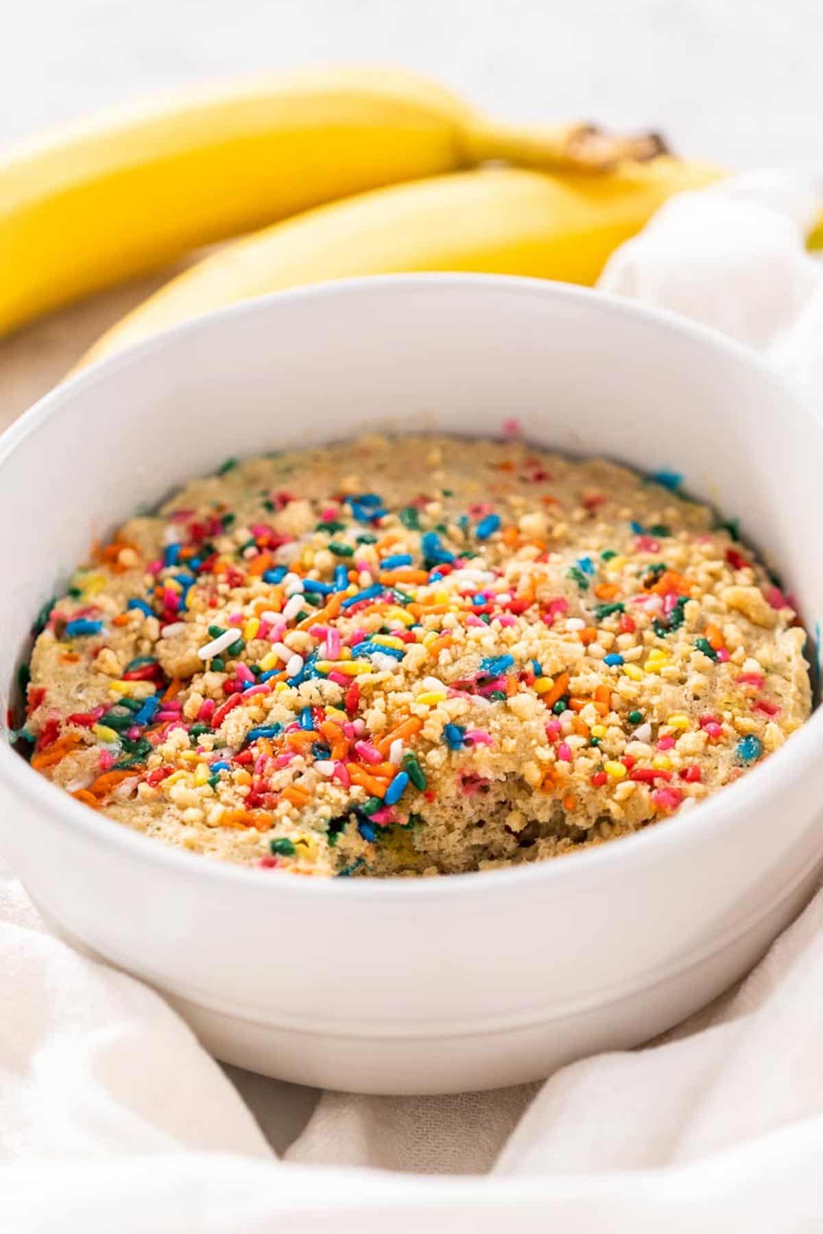 A white bowl of Baked Birthday Cake Oatmeal sitting on a white dish towel with two bananas in the background.