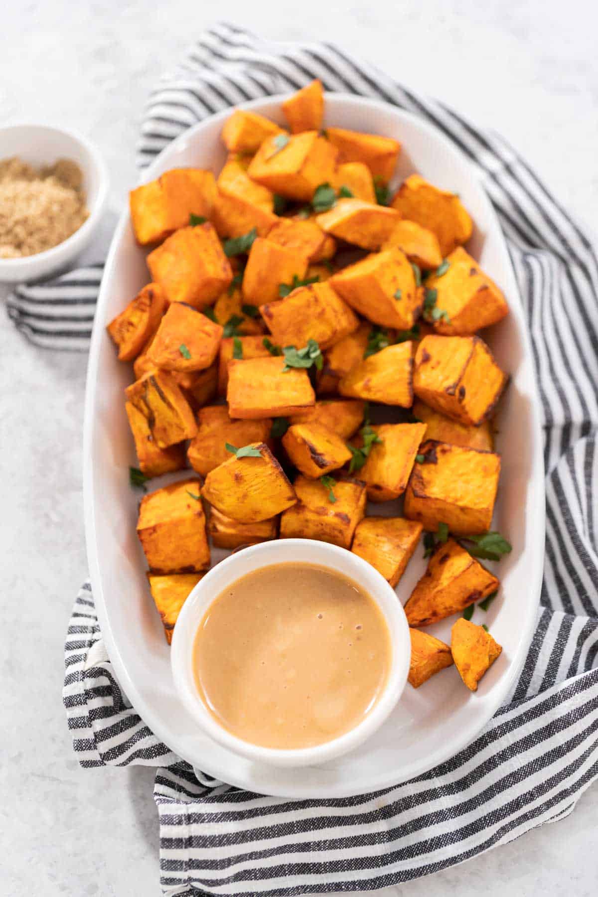 Brown sugar air fryer sweet potato cubes on a white plate, sauce in a white bowl and both are sitting upon a grey and white striped towel.