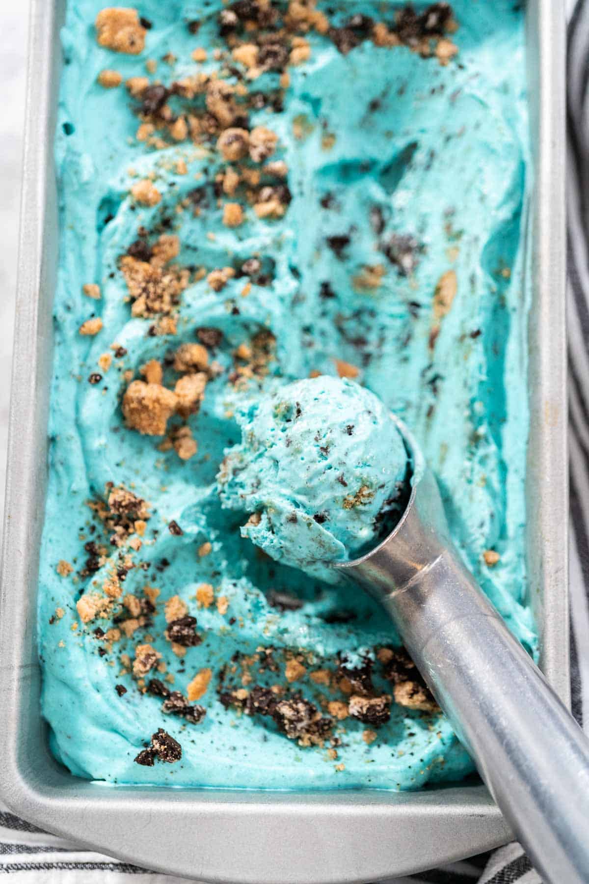 Cookie Monster Ice Cream, a blue ice cream with cookies crumbled on top in a metal pan and a metal ice cream scoop on top.