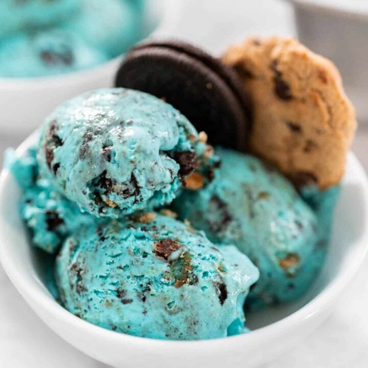 Cookie Monster Ice Cream in a white bowl with three scoops of blue ice cream with Oreos and cookies mixed in. A whole Oreo and cookie are placed in the bowl.