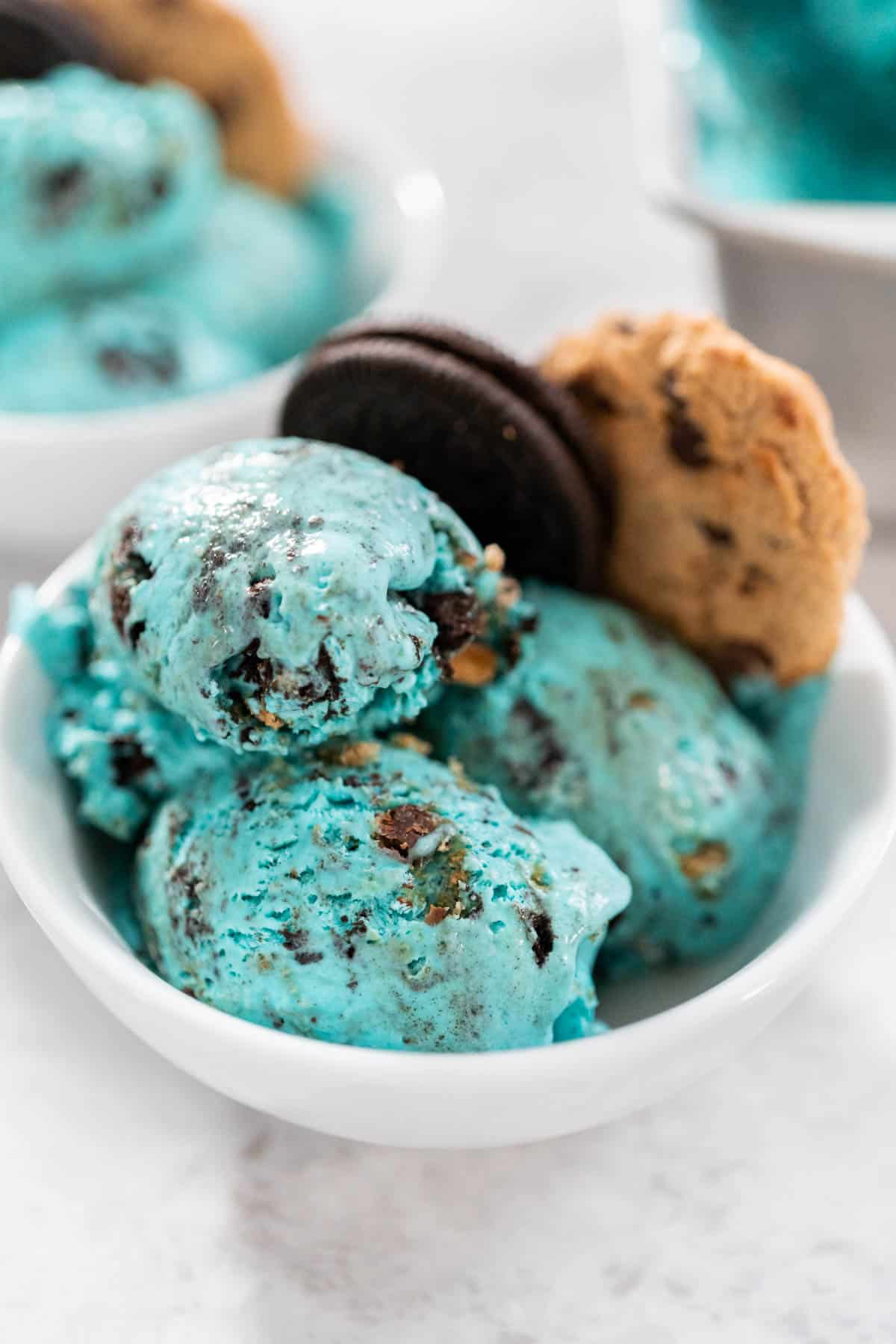 Cookie Monster Ice Cream in a white bowl with three scoops of blue ice cream with Oreos and cookies mixed in. A whole Oreo and cookie are placed in the bowl.