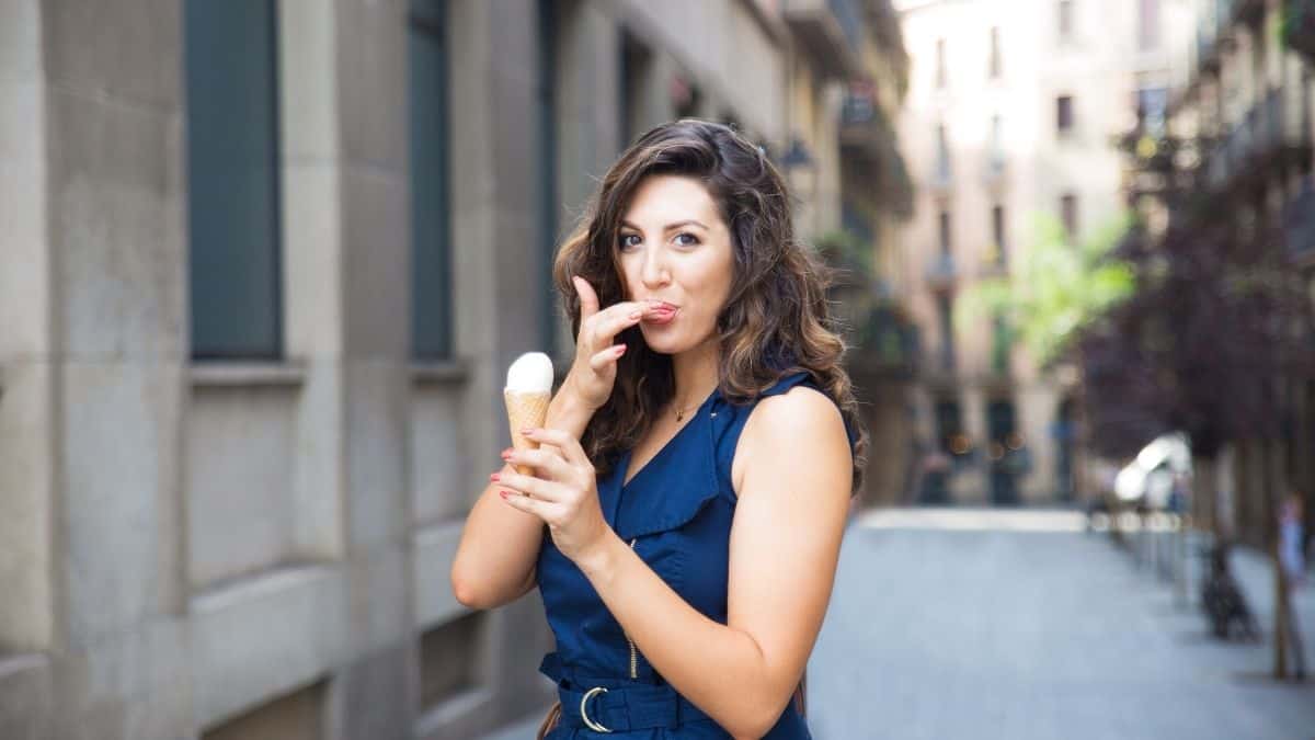 a brunette woman in a blue dress eating a vanilla ice cream cone and licking her finger.