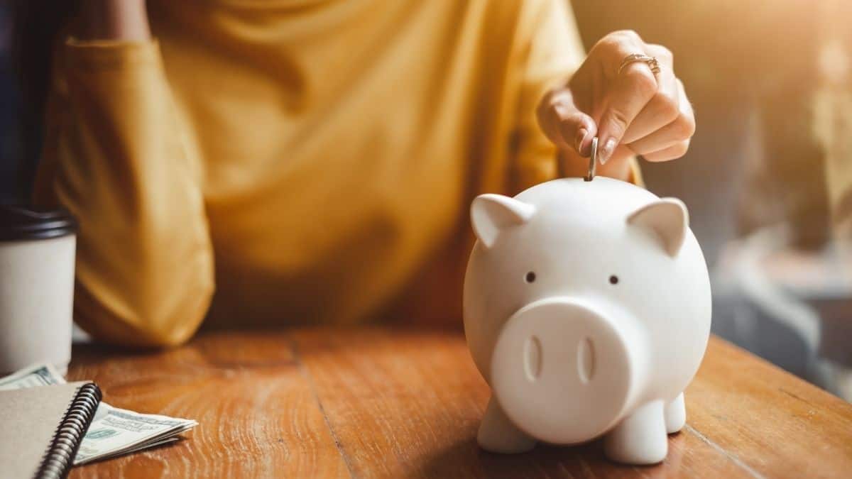 a woman wearing a mustard colored sweater puts a coin into a white piggy bank, representing money earned by the diet industry.