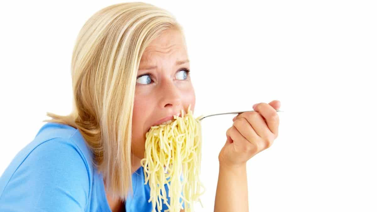 a blonde woman eating a large forkful of spaghetti noodles with a worried look on her face, an example of what a binge could look like.