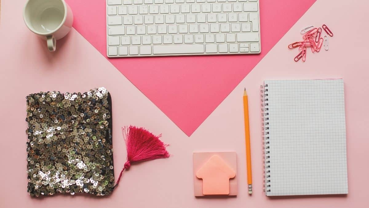 A computer, coffee cup, notepad and pencil on pink background.