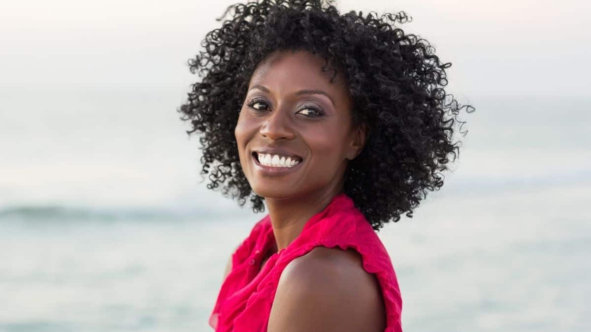 A woman with curly hair wearing a pink shirt with a joyful smile on her face.