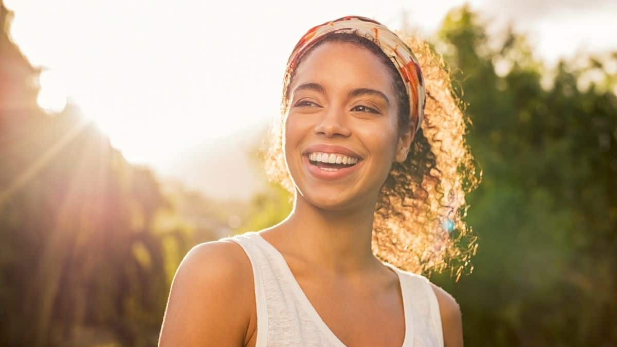 A woman with curly hair and big smile on her face in front of trees and the sun shining behind her.