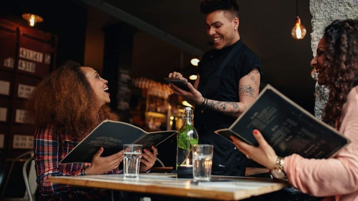 two women and a server at a restaurant smiling, clearly enjoying eating out.