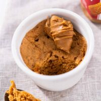 A scoop of biscoff mug cake on a metal spoon resting beside a white ramekin with mug cake and cookie butter on top.
