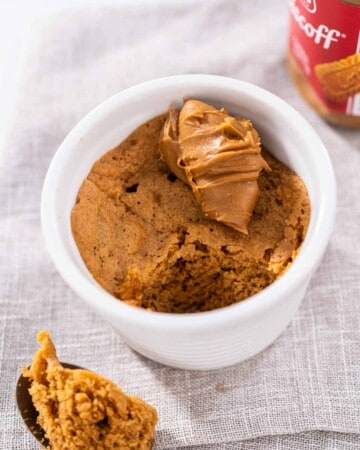 A scoop of biscoff mug cake on a metal spoon resting beside a white ramekin with mug cake and cookie butter on top.