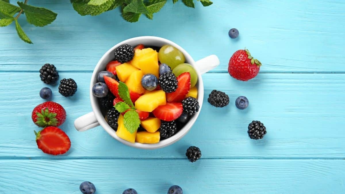 a white bowl of mixed fruit including peaches, blackberries, strawberries, blueberries with mint garnished on top on a blue background.