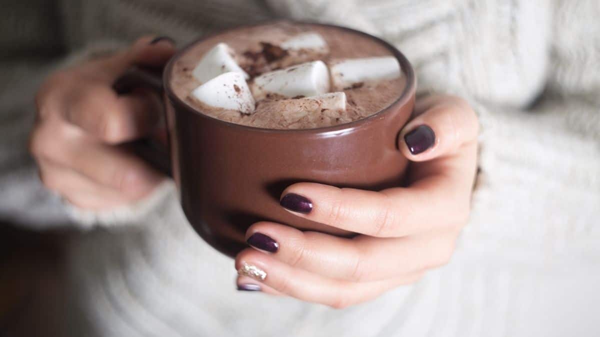 a woman's hands wrapped around a warm mug of hot chocolate topped with marshmallows, something someone may indulge in during the holiday season.