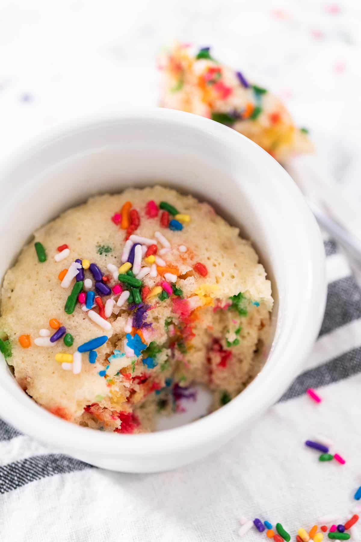 vanilla vegan mug cake in a white ramekin with a spoonful missing with sprinkles scattered around.