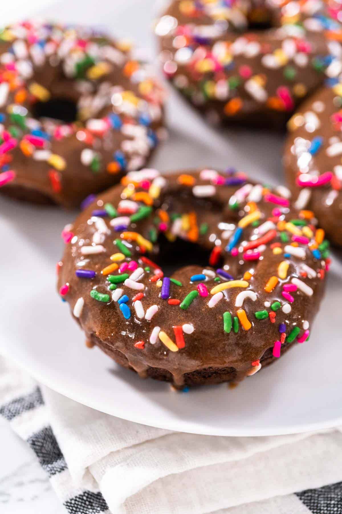 A close up of four chocolate protein donuts covered in rainbow sprinkles on a white plate.