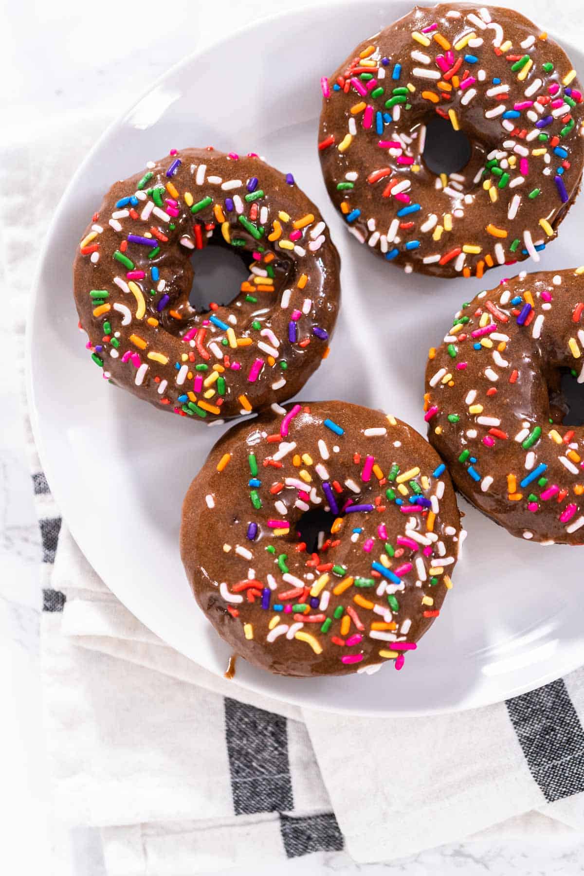 An overhead shot of four chocolate protein donuts covered in rainbow sprinkles on a white plate.