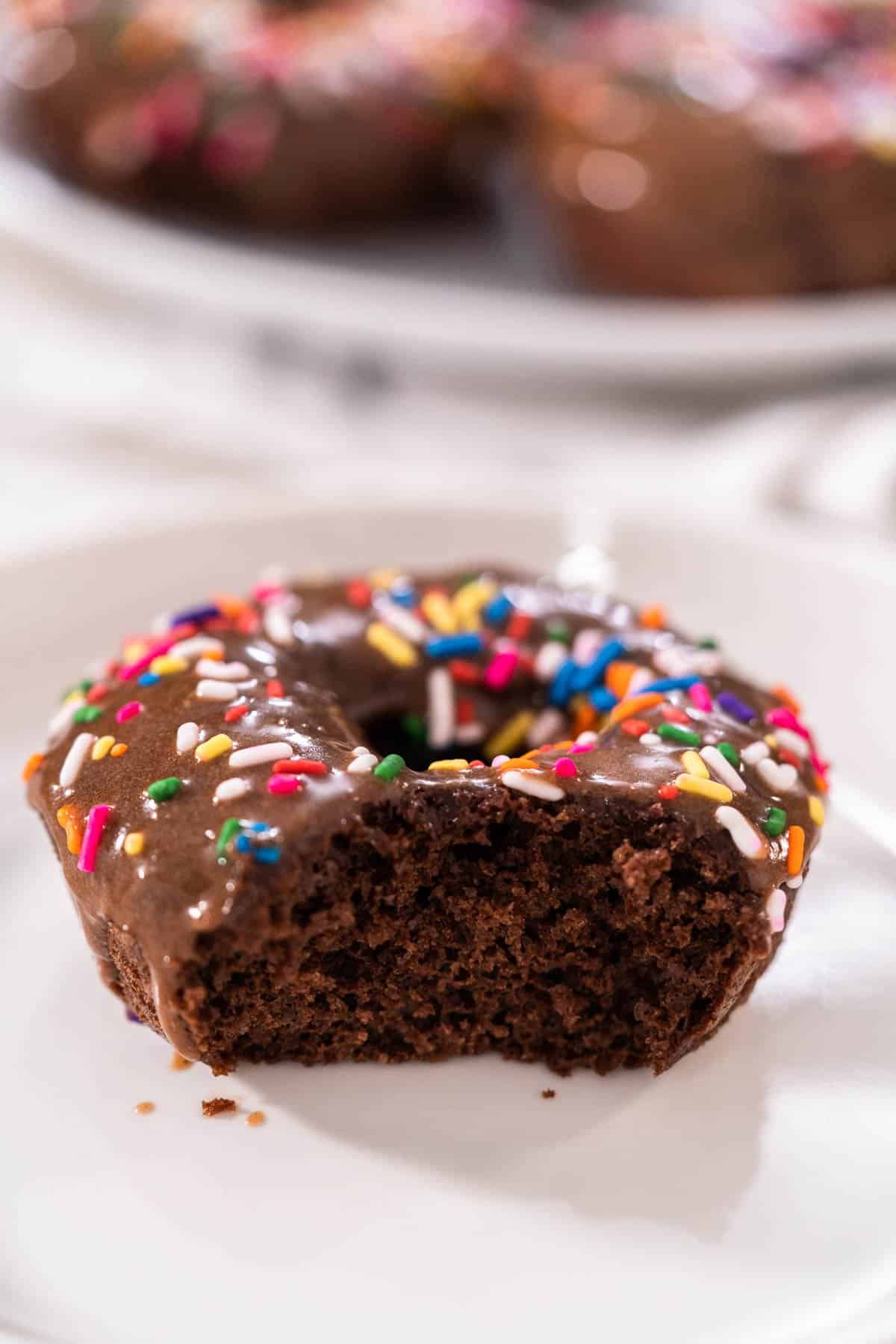 One chocolate protein donut covered in rainbow sprinkles with a bite taken out of it on a white plate.