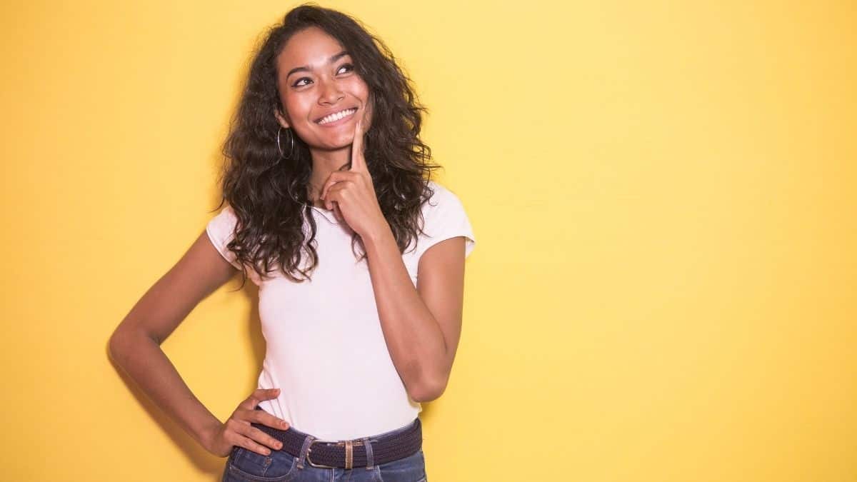 a smiling woman in a light pink tshirt with a finger to her chin contemplating something.