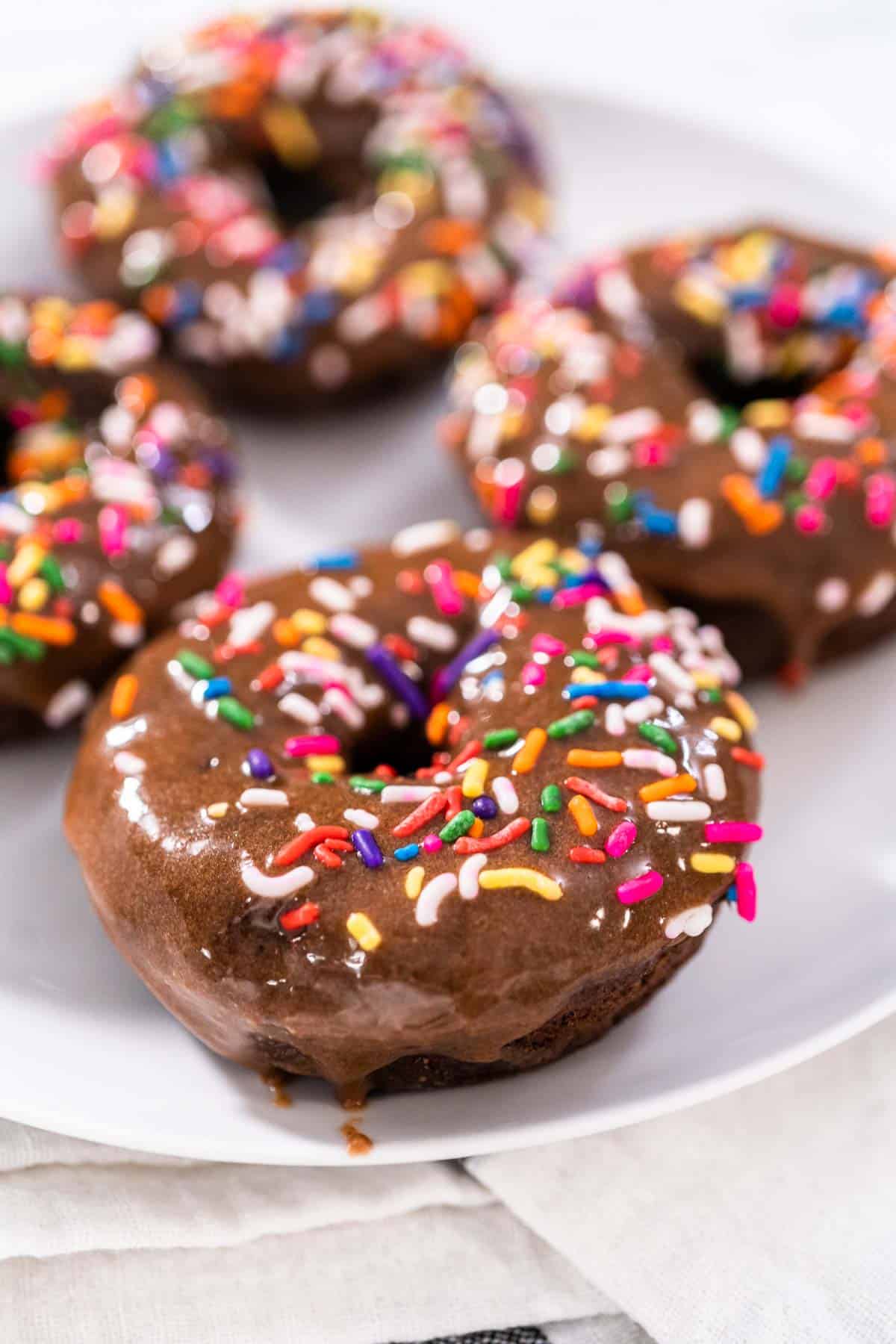 four chocolate protein donuts covered in rainbow sprinkles on a white plate.