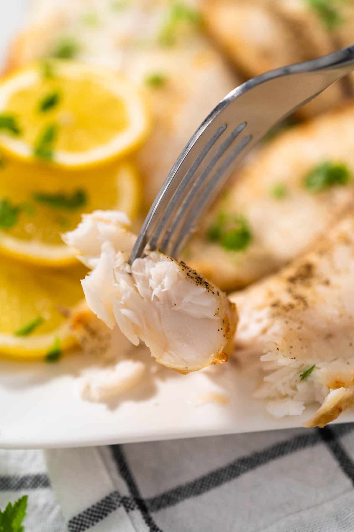 A close up of a bite of air fryer tilapia with a plate of tilapia and lemon slices in the background.