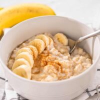 A white bowl of banana porridge with a sliced banana, maple and cinnamon on top.