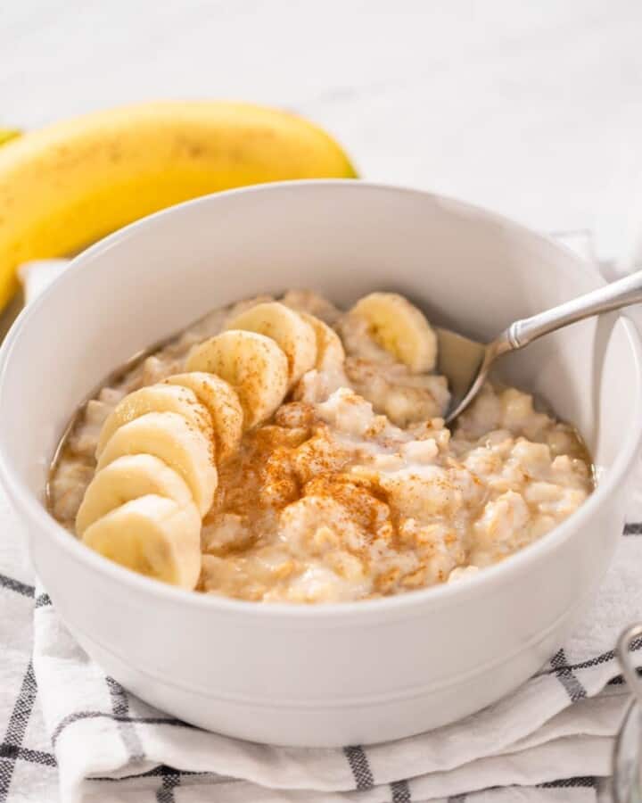 A white bowl of banana porridge with a sliced banana, maple and cinnamon on top.