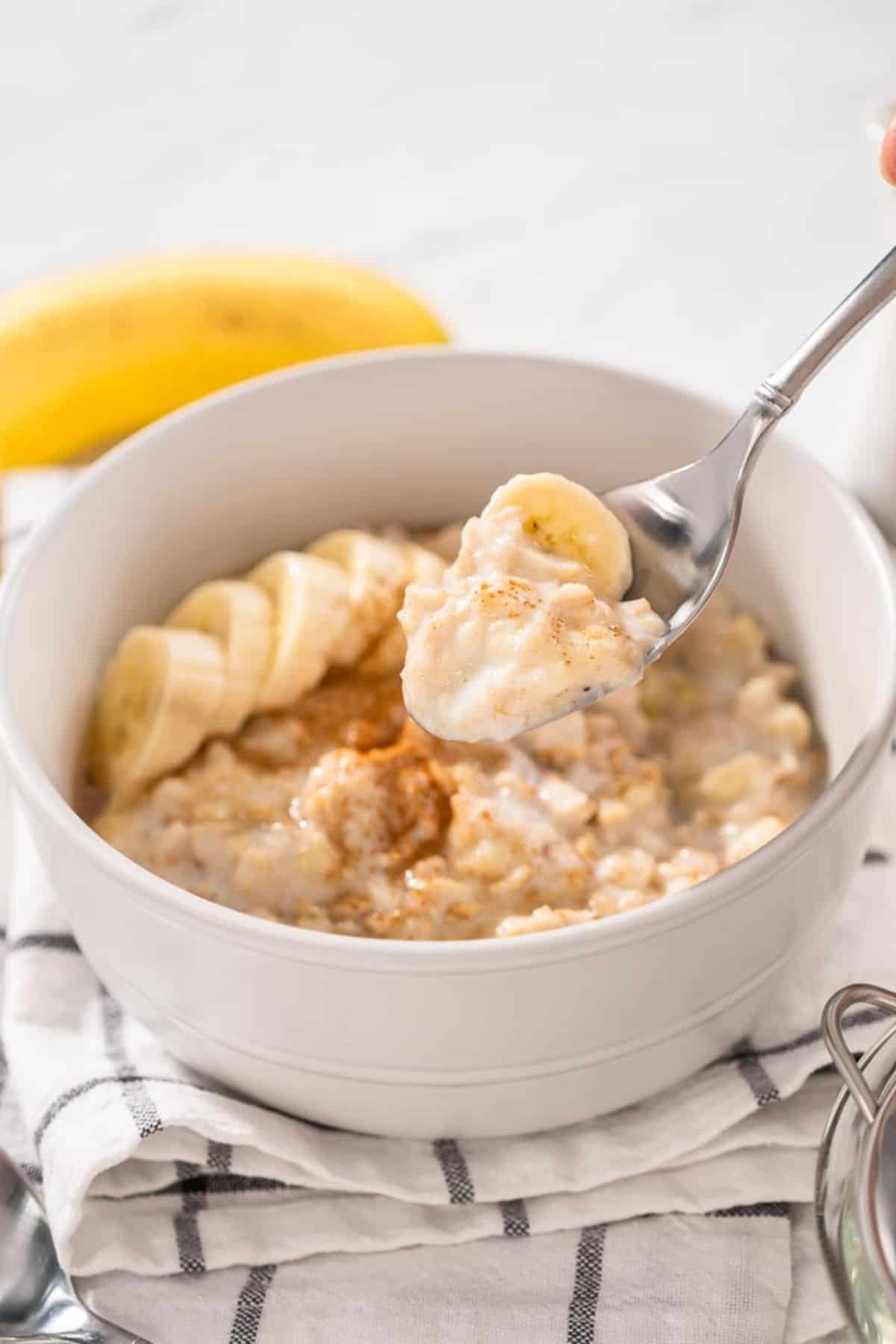 A woman taking a spoonful of banana porridge topped with banana from a white bowl.