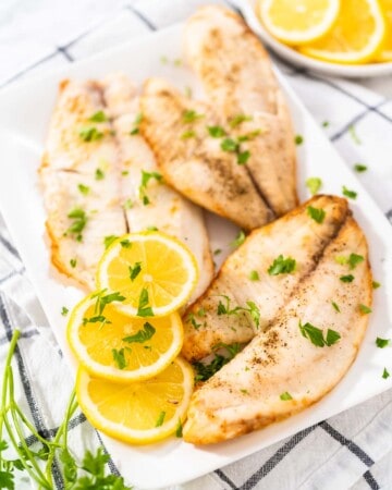 Three pieces of air fryer tilapia on a white plate with lemon slices.
